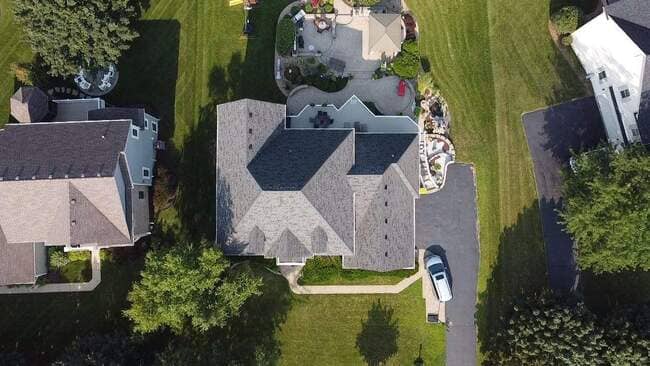 Aerial view of a newly installed dark gray asphalt shingle roof on a suburban house. The image captures the home's multi-gabled roofline, surrounding well-manicured lawn, and a back patio area with seating.